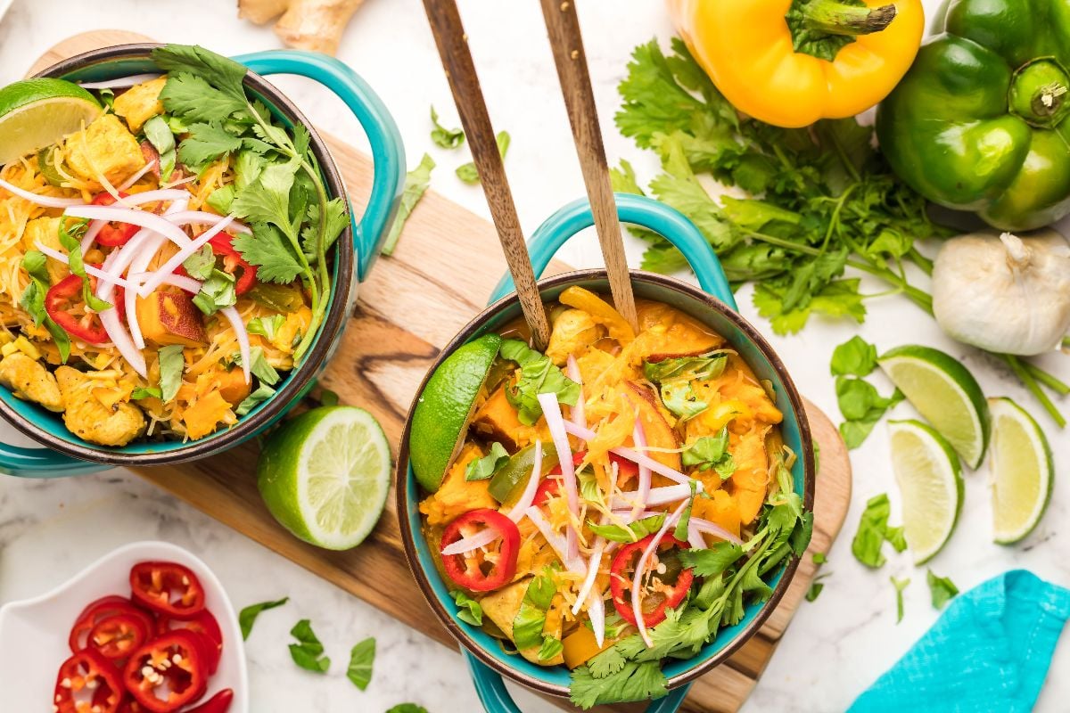 Two bowls of hearty Thai recipe with chicken and vegetables, garnished with cilantro, lime, and red chili, sit next to fresh peppers.