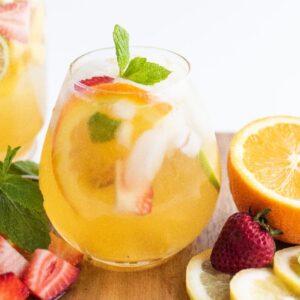 A glass of white sangria with summer fruit, infused with citrus, berries, ice, mint, and fruit slices served on a wooden board.