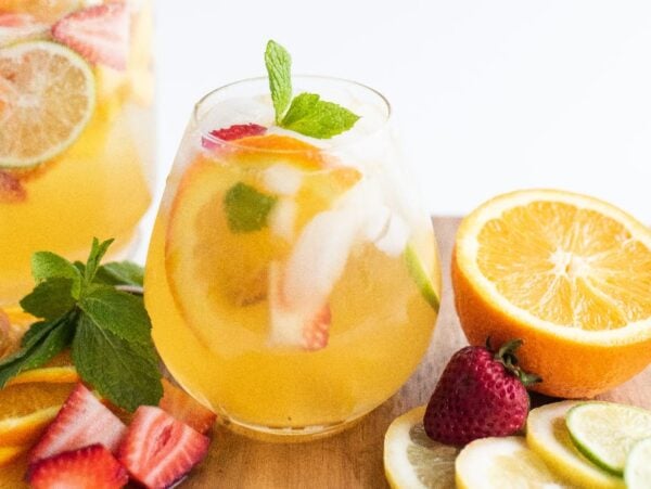 A glass of white sangria with summer fruit, infused with citrus, berries, ice, mint, and fruit slices served on a wooden board.