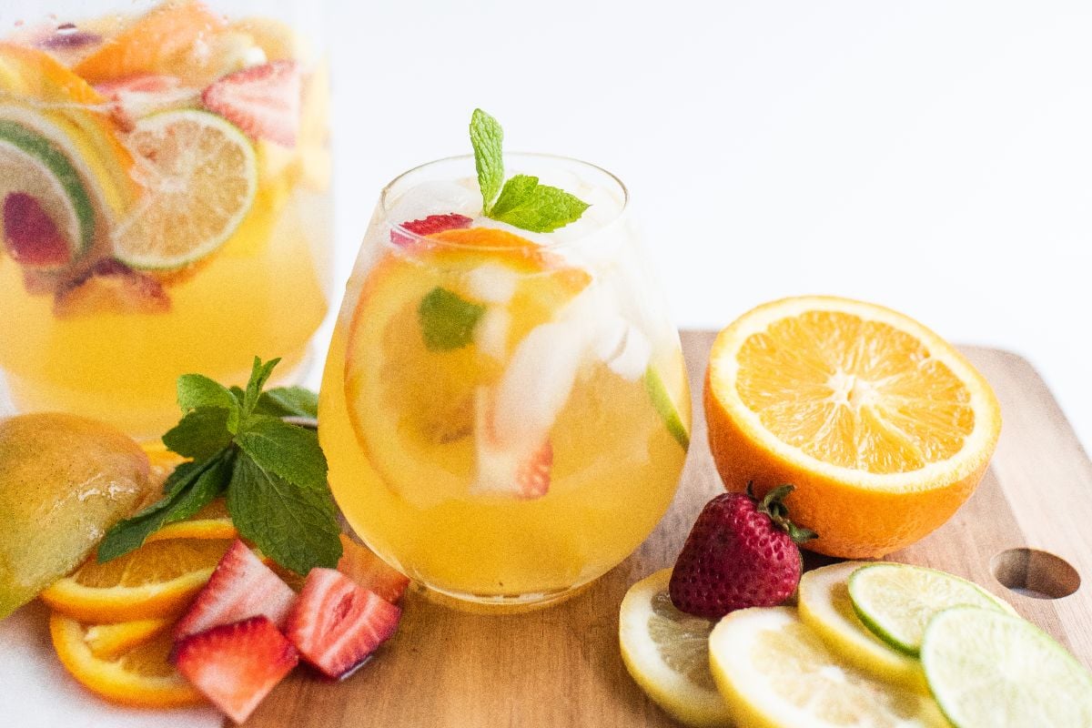 Glass of fruity iced drink with sliced lime, orange, and strawberries on a wooden board with fresh fruit—a super easy sangria recipe perfect as a refreshing summer drink.