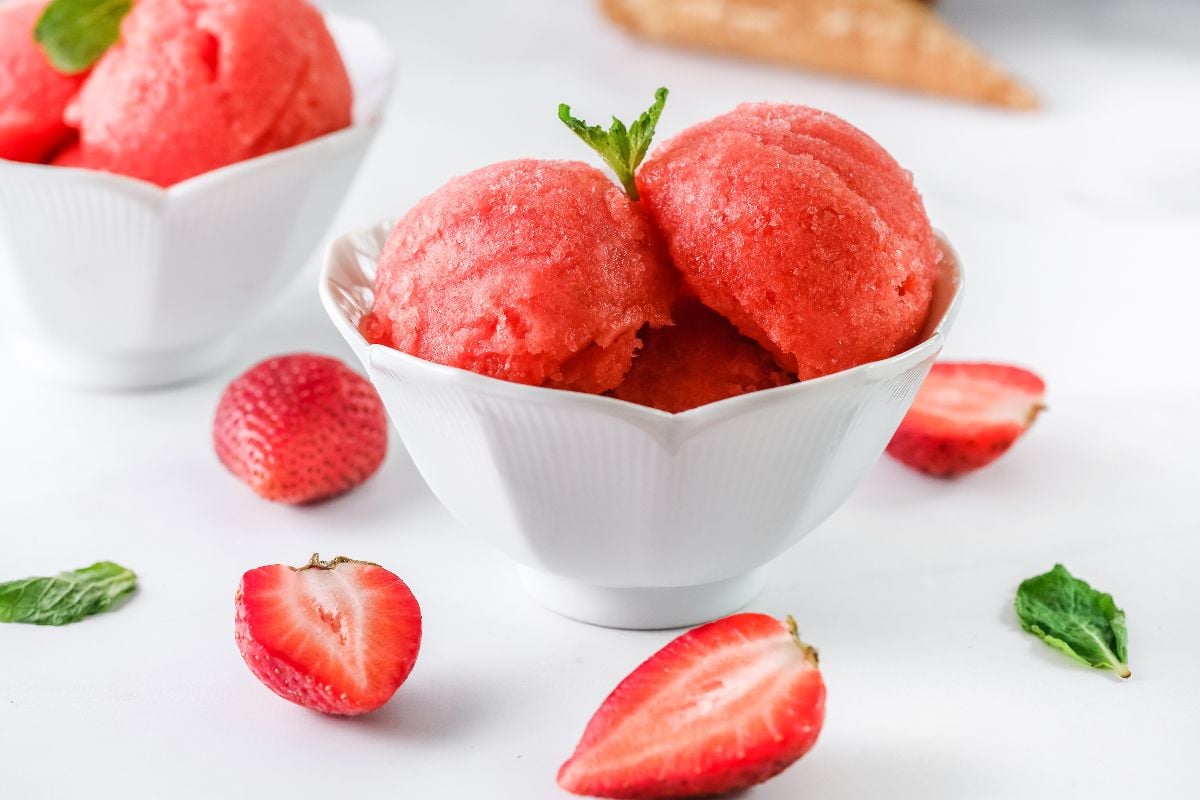 White bowl with scoops of strawberry sorbet, made with frozen strawberries and just 4 ingredients, garnished with mint—surrounded by fresh strawberries. A refreshing summer dessert idea!.