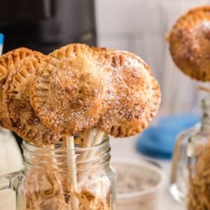 Mini s’mores pie pops dusted with powdered sugar in mason jars, with milk in bottles and striped straws nearby—a sweet twist inspired by the air fryer s’more recipe.