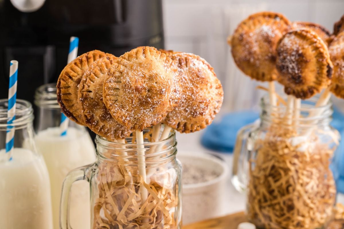 Round pie pops on sticks in mason jars, with milk in glass jars and striped straws in the background—an easy s’more treat and perfect summer BBQ dessert idea.