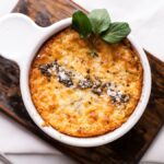 Golden baked dish in a white ramekin, garnished with herbs and black pepper, on a wooden board—a delicious hot artichoke appetizer perfect as an easy appetizer dip recipe.
