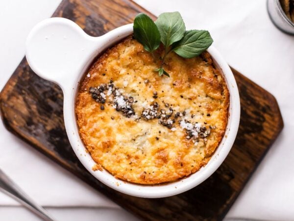 Golden baked dish in a white ramekin, garnished with herbs and black pepper, on a wooden board—a delicious hot artichoke appetizer perfect as an easy appetizer dip recipe.