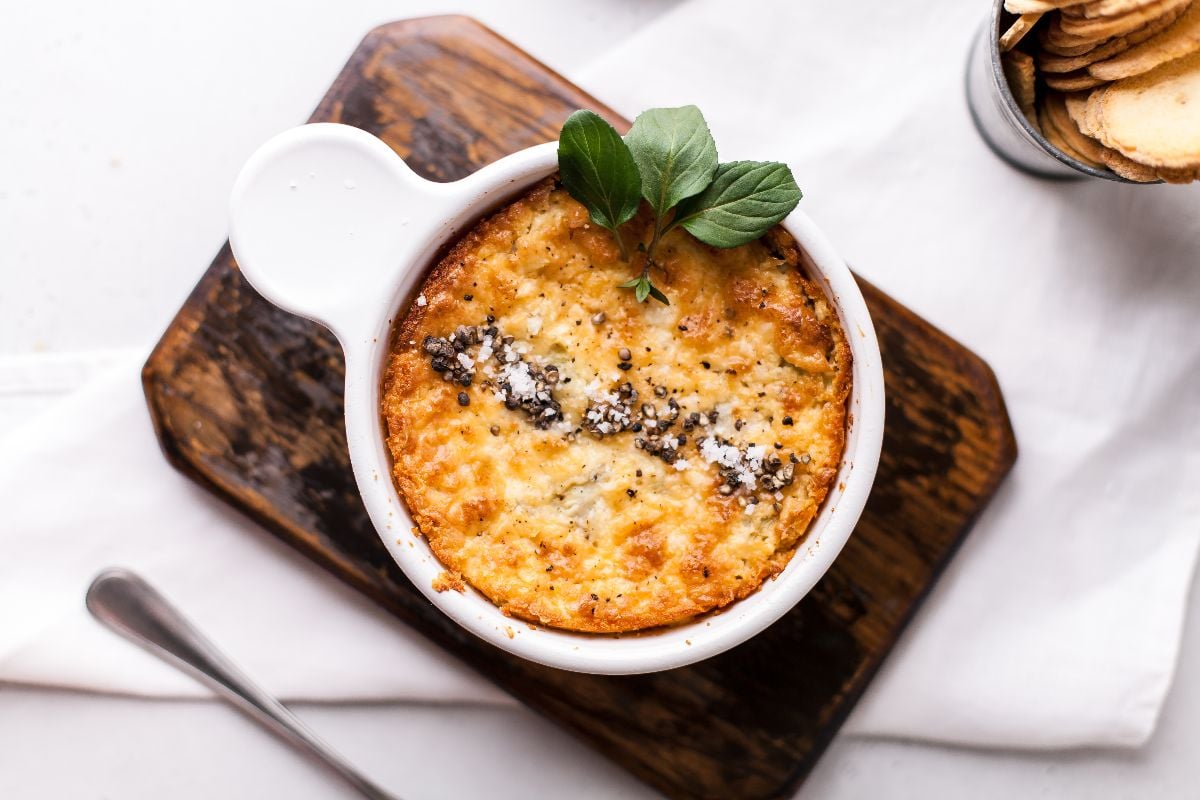 Baked artichoke dip in a white ramekin, garnished with herbs, on a wooden board with a spoon and crackers nearby—an easy appetizer dip recipe perfect for any gathering.