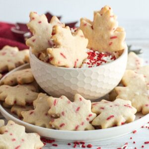 Maple leaf-shaped cookies with red sprinkles, perfect as Canada Day cookies, are displayed on a white plate and in a bowl on a white table.