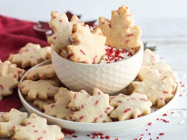 Maple leaf-shaped cookies with red sprinkles, perfect as Canada Day cookies, are displayed on a white plate and in a bowl on a white table.