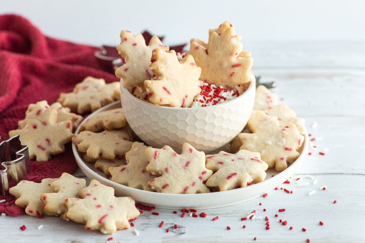 A plate of maple leaf-shaped cookies with red sprinkles, some stacked in a bowl, on a white surface—perfect cookies to celebrate Canada Day or inspire your next Canada Day dessert ideas.
