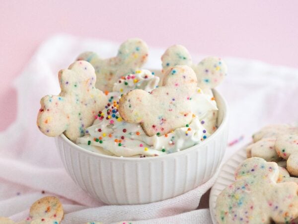 Bowl of funfetti Dunkaroo dip with colorful sprinkle cookies shaped like teddy bears on a pink background.