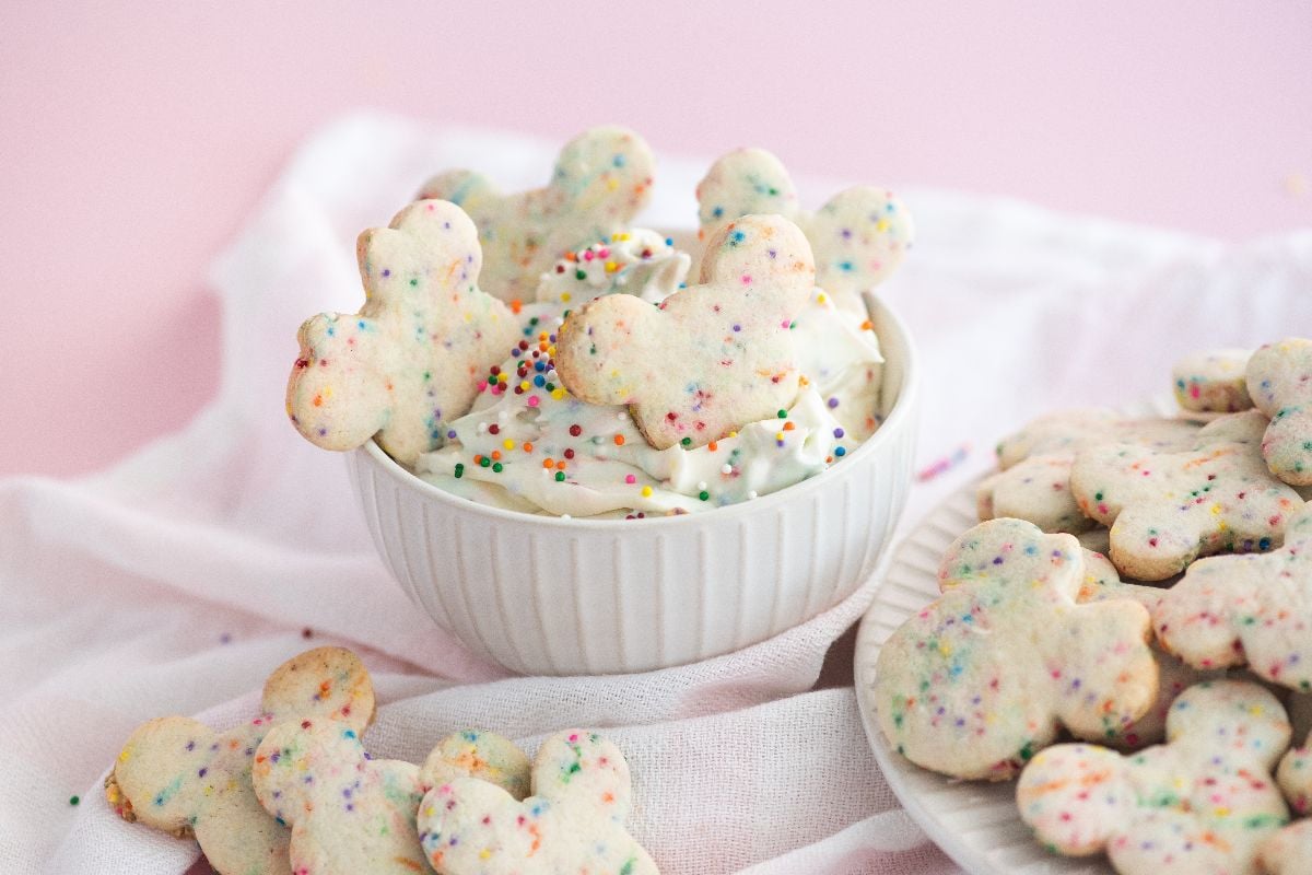 Bowl of cream cheese sprinkle dip with Mickey-shaped Disney Sugar Cookies with Dip, some dipped, on a soft white cloth.