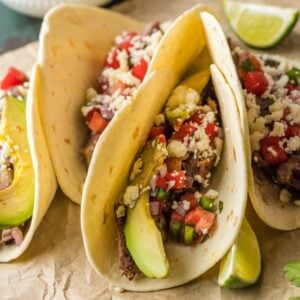 Three tacos filled with grilled meat using the best flank steak marinade, avocado, pico de gallo, cheese, and lime wedges on the side.