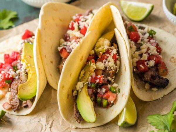 Three tacos filled with grilled meat using the best flank steak marinade, avocado, pico de gallo, cheese, and lime wedges on the side.