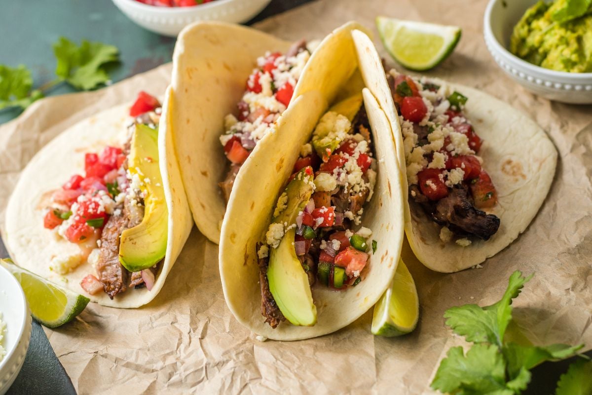 Three soft tacos filled with savory grilled meat, avocado, pico de gallo, and cheese on parchment paper—perfect for enjoying classic flank steak tacos.