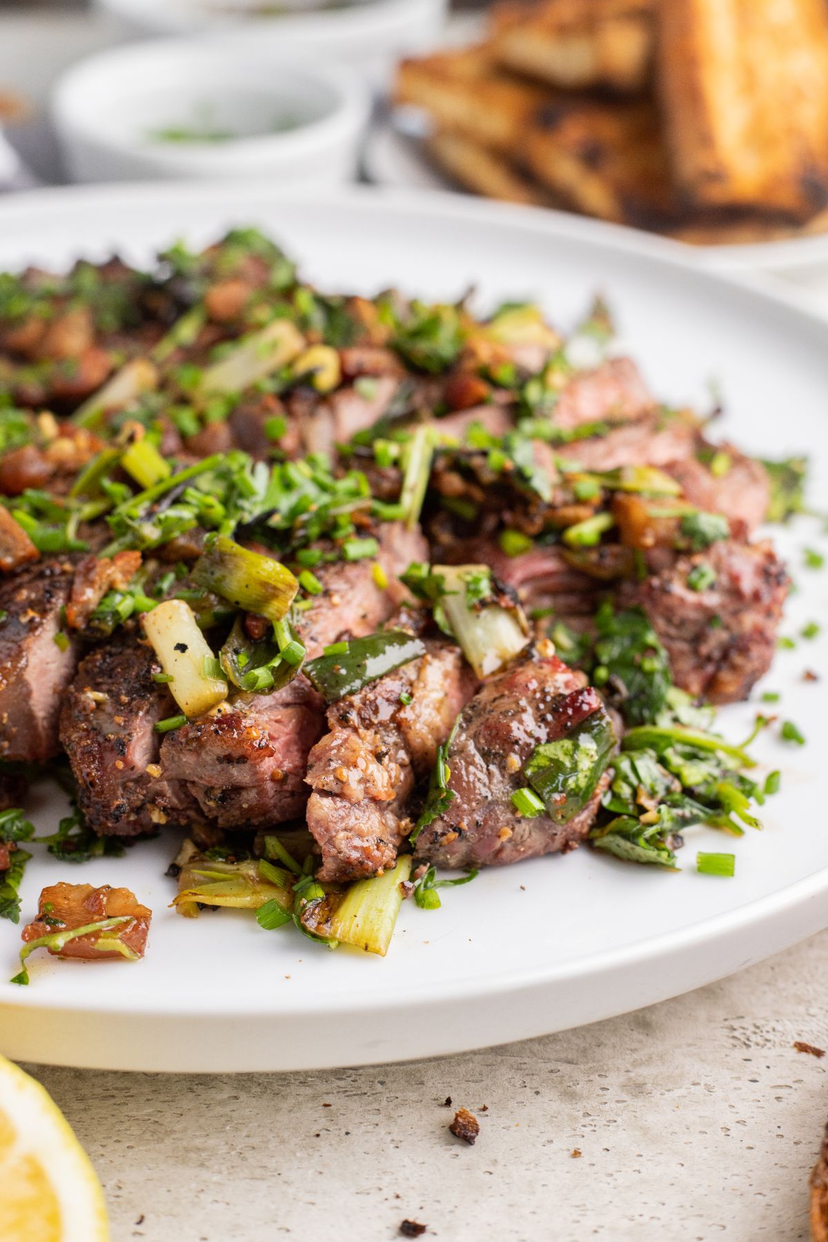 Sliced grilled steak with herbed scallion topping, finished with chopped herbs and vegetables on a white plate.