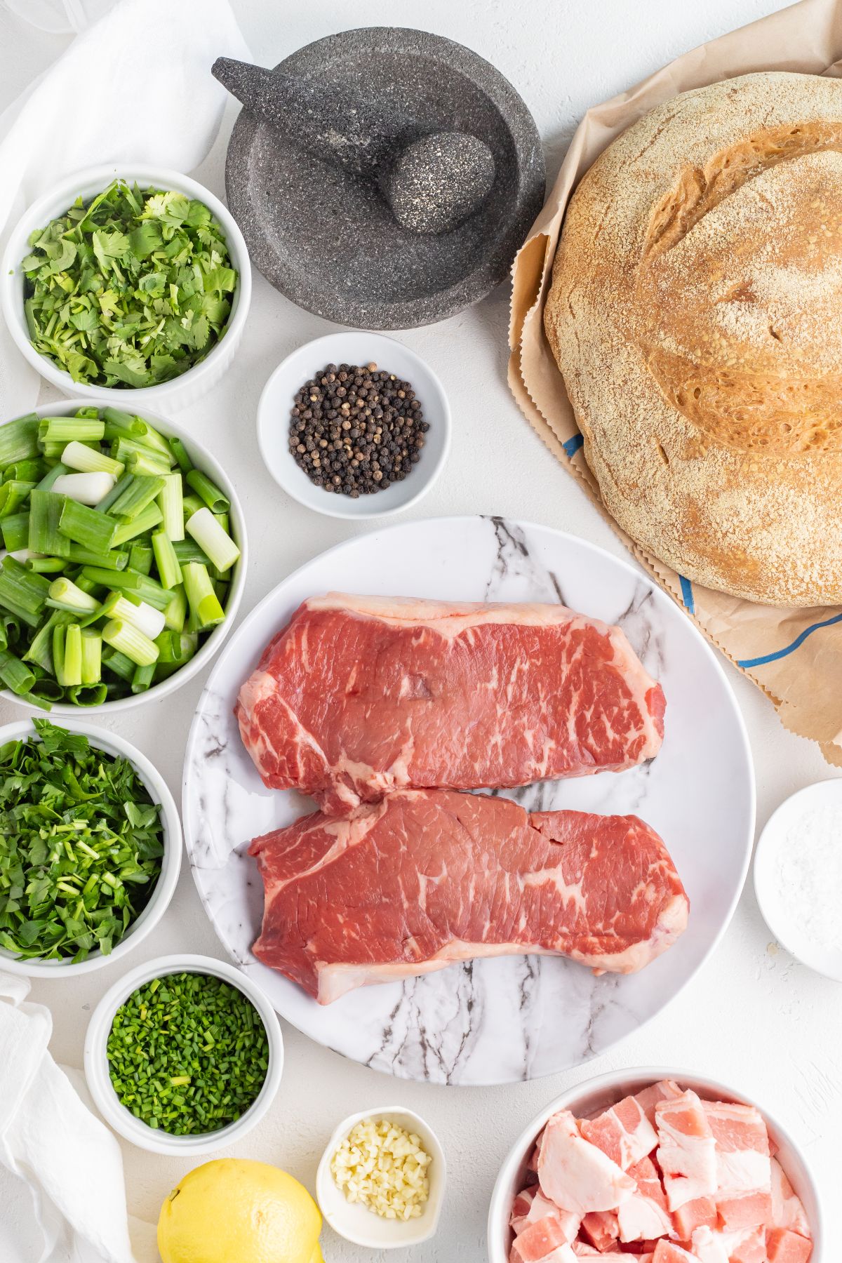 Raw steaks on a plate, surrounded by chopped herbs, green onions, bread, spices, lemon, and diced pork—perfect for trying a new BBQ steak recipe or serving as Grilled Steak with Herbed Scallion Topping.