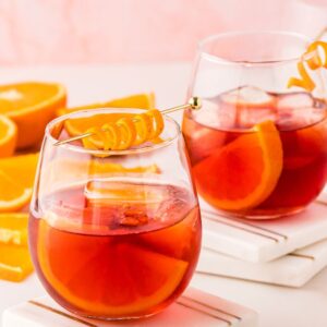 Two glasses of negroni cocktail with ice and orange garnishes, surrounded by sliced oranges on a white surface—a classic Italian cocktail perfect for learning how to make the best negroni at home.
