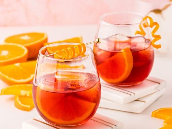 Two glasses of negroni cocktail with ice and orange garnishes, surrounded by sliced oranges on a white surface—a classic Italian cocktail perfect for learning how to make the best negroni at home.