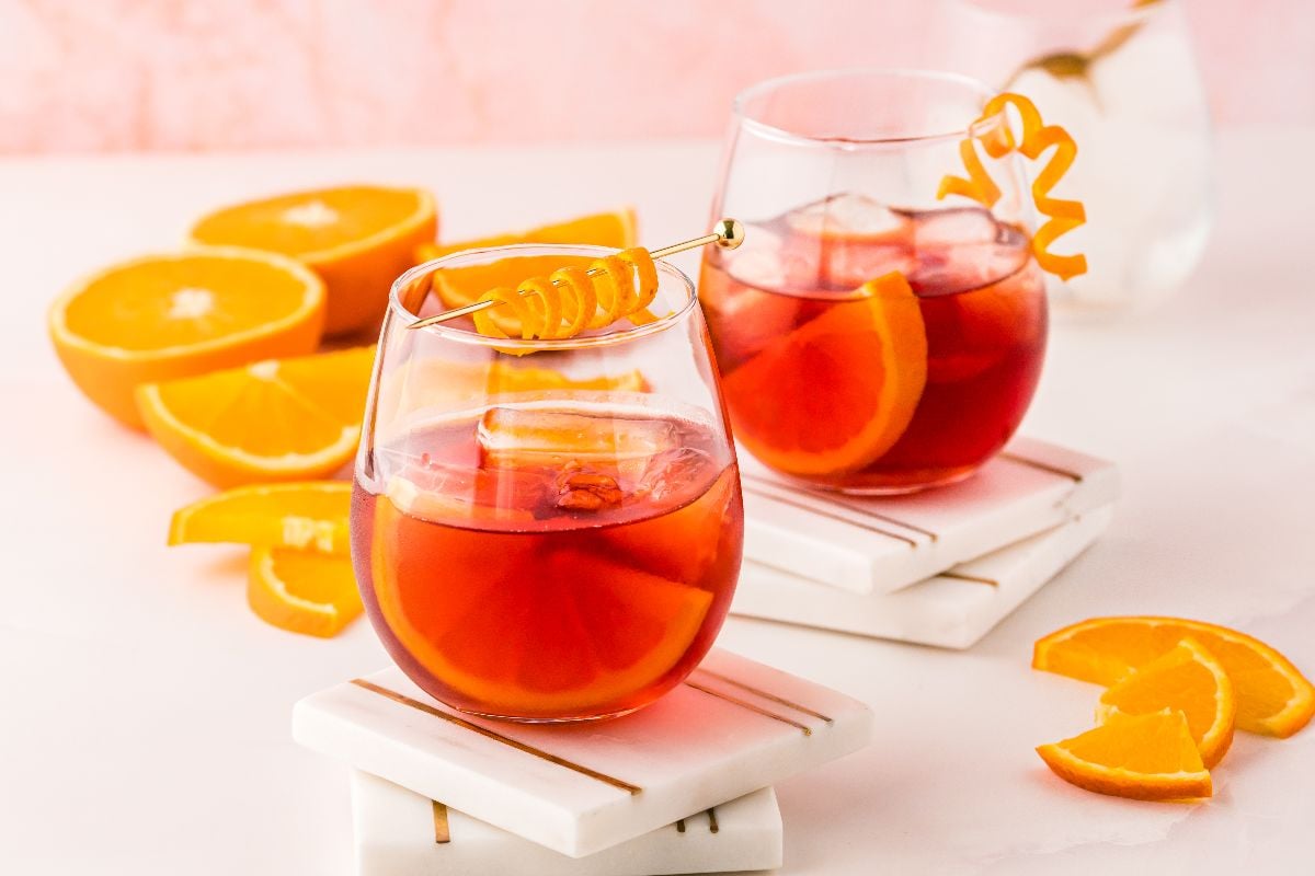 Two glasses of iced negroni cocktail with orange slices and garnish, surrounded by orange wedges on a white surface.