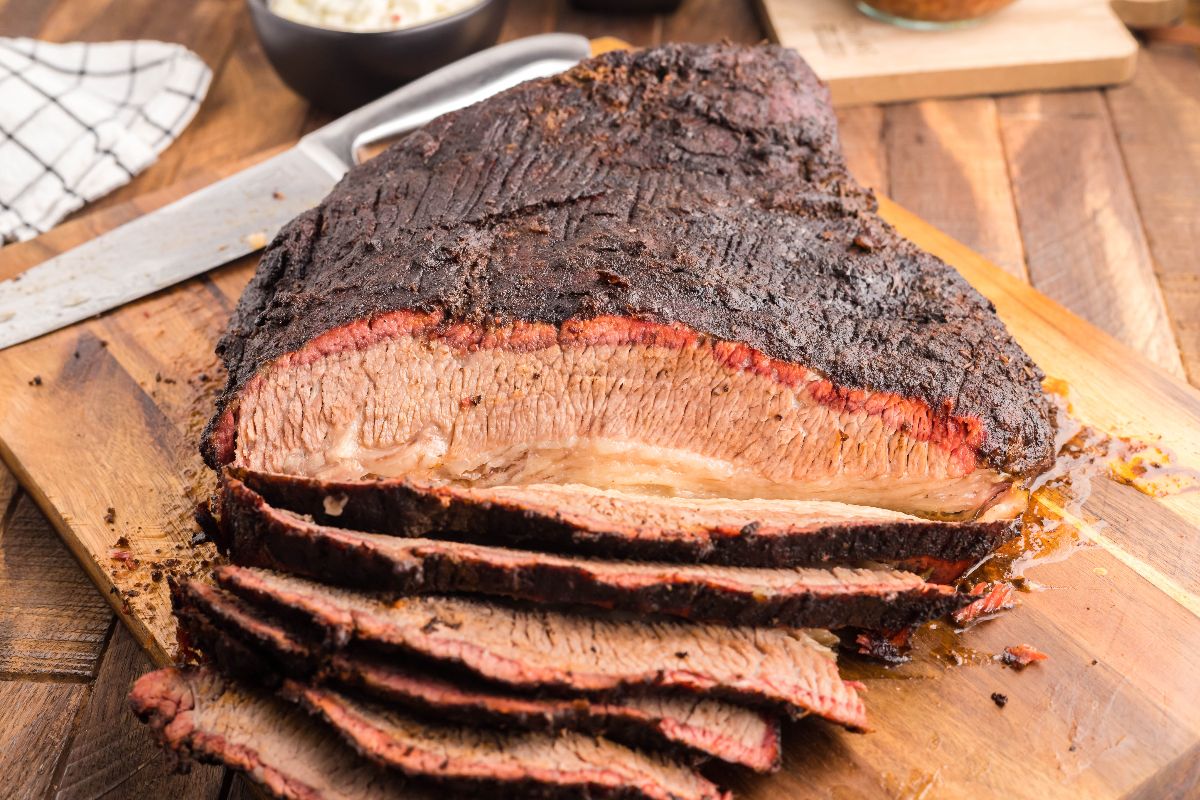 Sliced smoked beef brisket on a wooden cutting board with a knife beside it, showcasing the perfect dry rub for brisket—a savory result of brisket made in a Traeger grill.