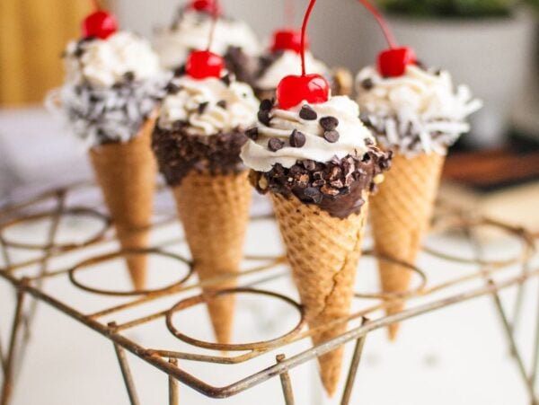 Mini waffle cones filled with creamy ricotta cannoli filling, whipped cream, chocolate, and cherries, displayed on a metal stand.