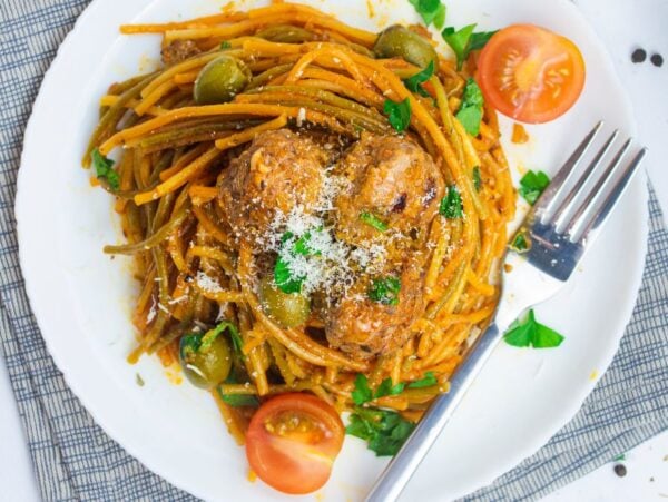 Plate of Mediterranean spaghetti and meatballs, topped with tomato sauce, olives, cherry tomatoes, parsley, and grated cheese.