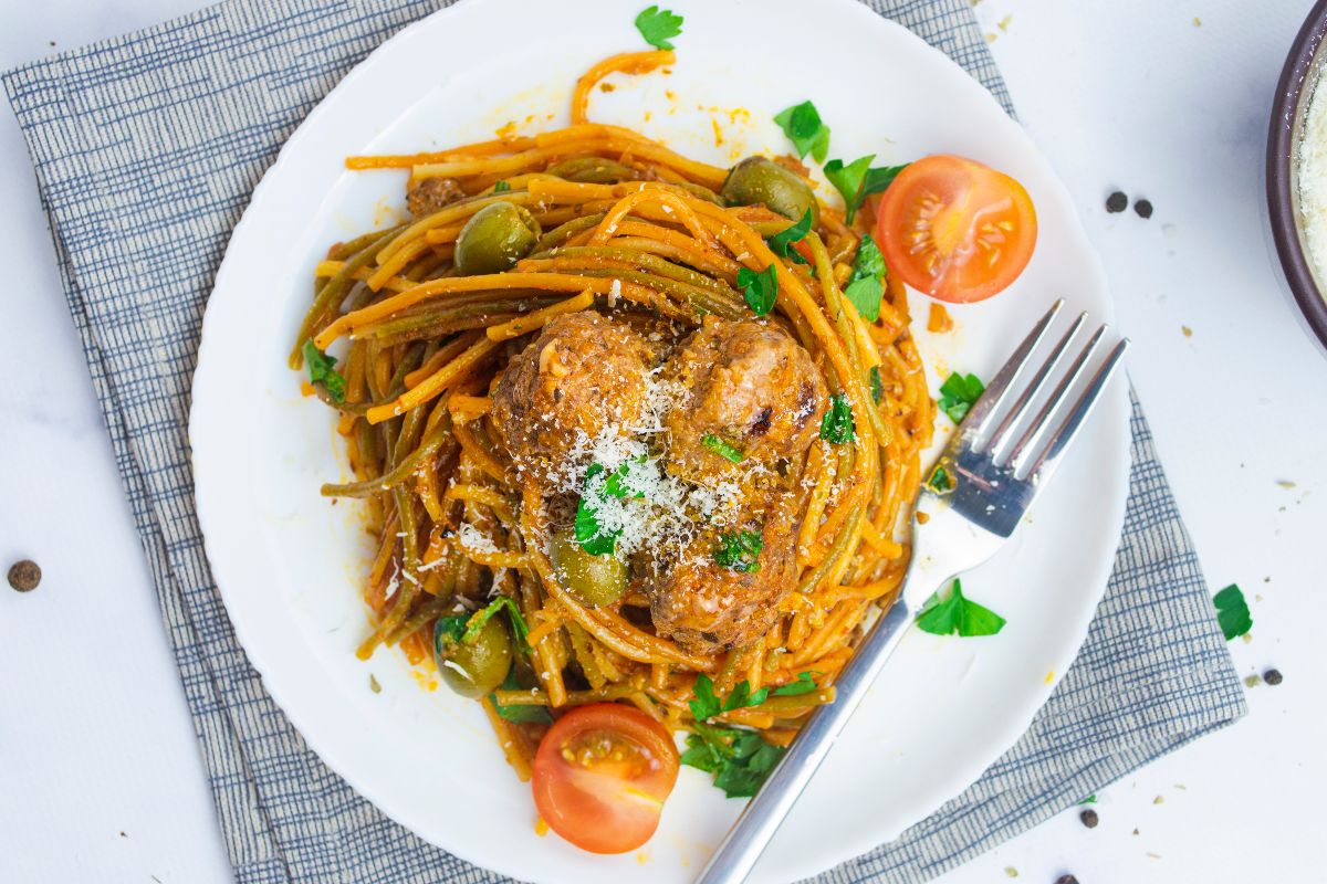 Enjoy this easy one pot Mediterranean pasta: spaghetti with meatballs, olives, cherry tomatoes, and parsley, all topped with grated cheese on a white plate.
