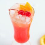 A tall glass of pink cocktail with ice, orange slices, and cherries, with orange slices on the side—perfect for those searching for an easy rum runner recipe.