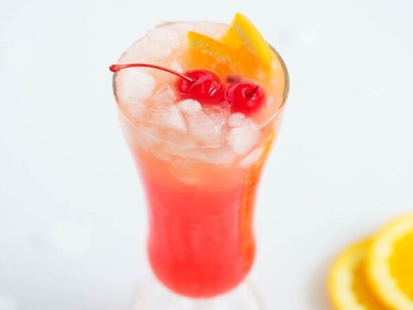 A tall glass of pink cocktail with ice, orange slices, and cherries, with orange slices on the side—perfect for those searching for an easy rum runner recipe.