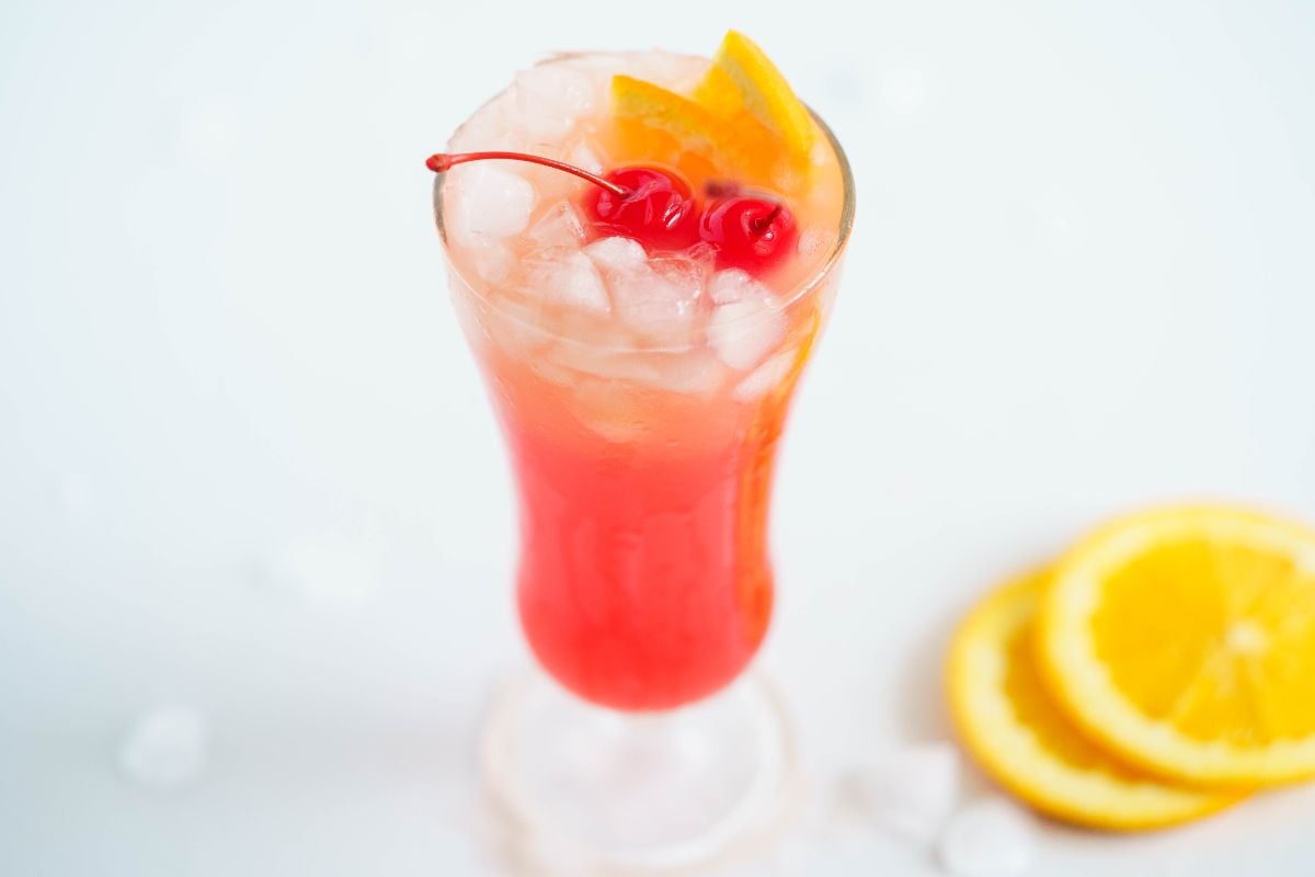 A tall glass of red-orange cocktail with ice, garnished with cherries and orange slices—perfect for trying an easy rum runner recipe or your favorite Caribbean punch cocktail recipe.