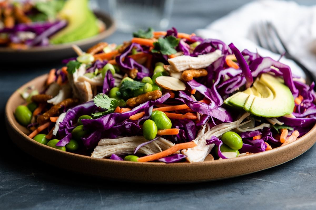 Colorful cabbage chicken salad with shredded chicken, purple cabbage, carrots, edamame, avocado, nuts, and cilantro on a plate, served with a tangy honey ginger dressing.