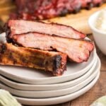 Three smoked ribs made in a Traeger grill are stacked on white plates next to a bowl of mashed potatoes on a wooden table—a Summer BBQ favourite.