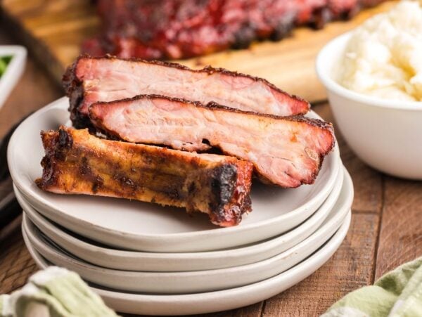 Three smoked ribs made in a Traeger grill are stacked on white plates next to a bowl of mashed potatoes on a wooden table—a Summer BBQ favourite.