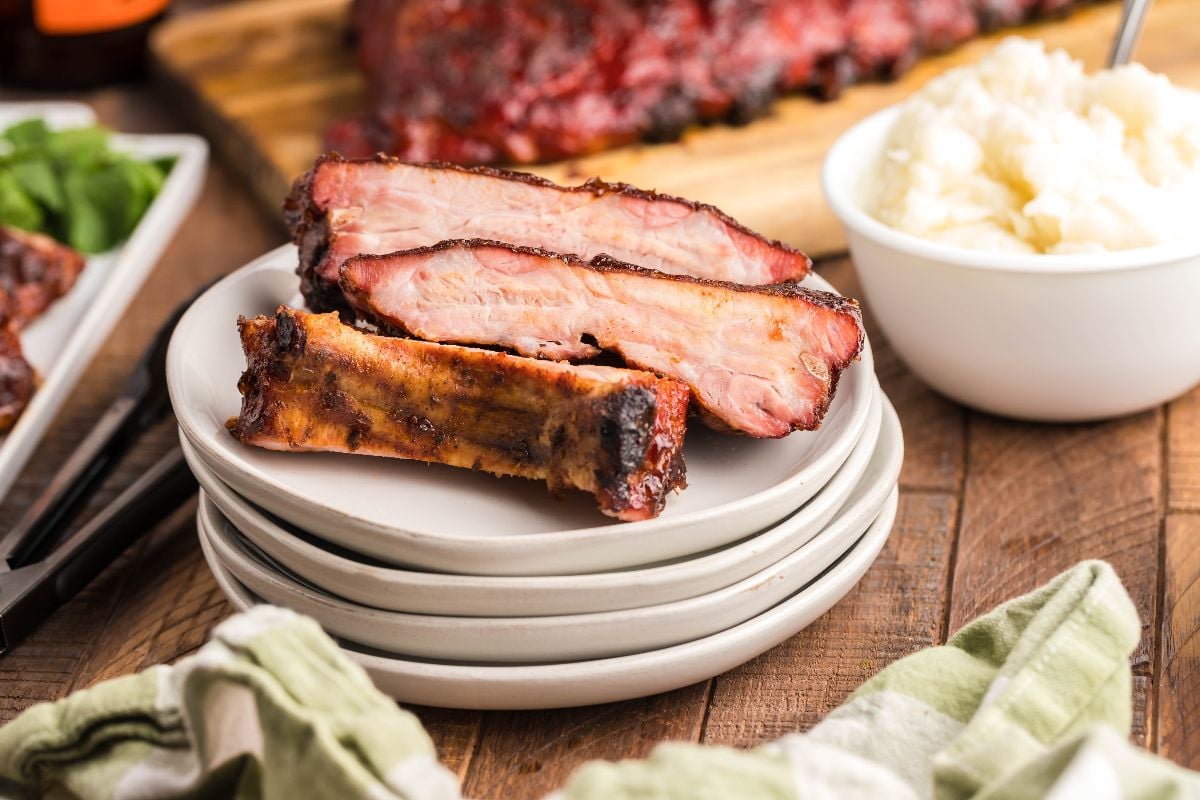 Three smoked baby back ribs stacked on white plates beside a bowl of mashed potatoes on a wooden table—an easy grilling recipe and a true Summer BBQ favourite.