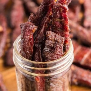 Thin strips of smoked beef jerky stand upright in a glass jar, with more healthy homemade beef jerky scattered on a wooden surface.