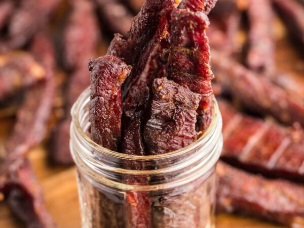 Thin strips of smoked beef jerky stand upright in a glass jar, with more healthy homemade beef jerky scattered on a wooden surface.