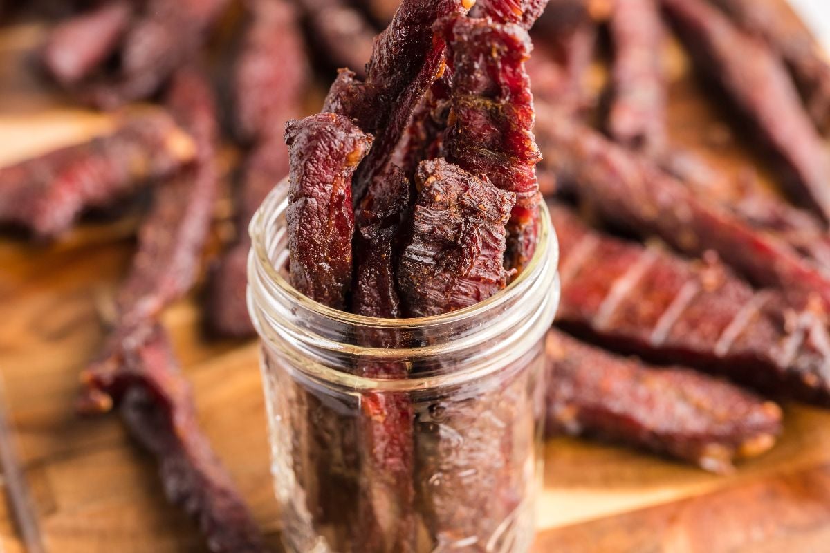 Several pieces of healthy homemade beef jerky stand upright in a glass jar, with more jerky on a wooden surface in the background—perfect inspiration if you're looking for a recipe for a smoker like a Traeger grill.
