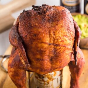 A roasted whole chicken sits upright on a beer can, ready for your next Traeger beer can chicken cookout, surrounded by cans and delicious sides on a wooden board.