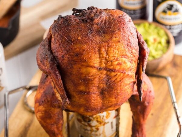 A roasted whole chicken sits upright on a beer can, ready for your next Traeger beer can chicken cookout, surrounded by cans and delicious sides on a wooden board.