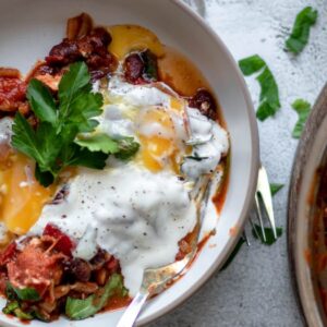 A bowl of shakshuka—eggs poached in a tomato sauce—topped with yogurt, fresh parsley, black pepper, and spinach for a savoury breakfast recipe.