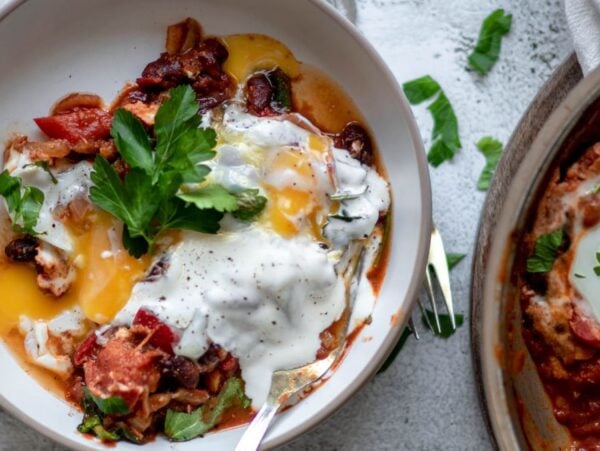 A bowl of shakshuka—eggs poached in a tomato sauce—topped with yogurt, fresh parsley, black pepper, and spinach for a savoury breakfast recipe.