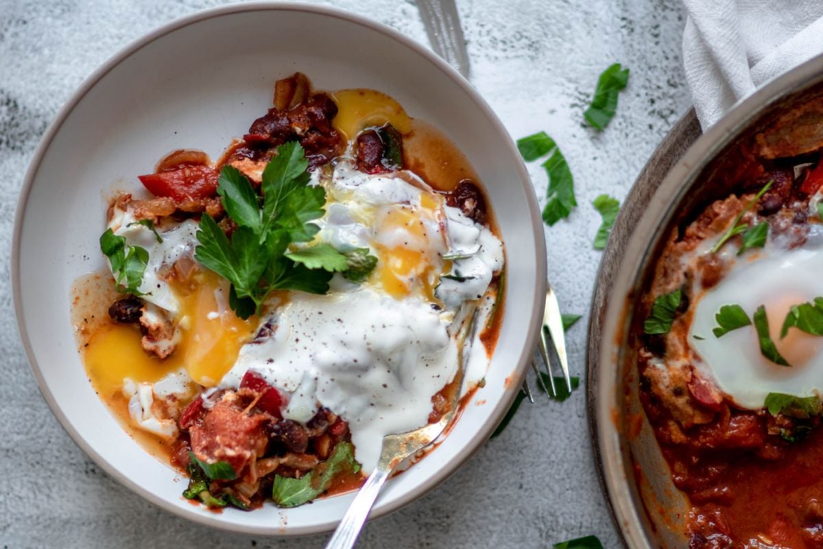 A bowl of spinach and red bean shakshuka topped with a runny egg, yogurt, and fresh parsley on a light textured surface—a quick healthy meal.