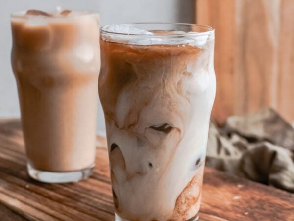 Two glasses of iced coffee with milk swirl, placed on a wooden surface with a beige background—perfect for making a Starbucks Copycat Iced Brown Sugar Oatmilk Shaken Espresso at home.