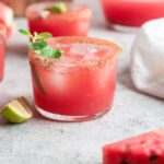 A glass of cocktail made with watermelon, ice, mint, and a chili rim, surrounded by lime wedges and watermelon slices—an easy summer cocktail recipe perfect for warm days.