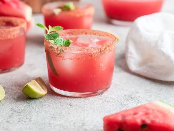 A glass of cocktail made with watermelon, ice, mint, and a chili rim, surrounded by lime wedges and watermelon slices—an easy summer cocktail recipe perfect for warm days.