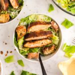 Bowl of Caesar salad topped with air fryer blackened chicken strips and croutons on a white surface, served with the best homemade Caesar dressing.