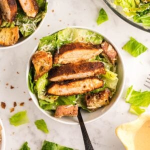 Bowl of Caesar salad topped with air fryer blackened chicken strips and croutons on a white surface, served with the best homemade Caesar dressing.