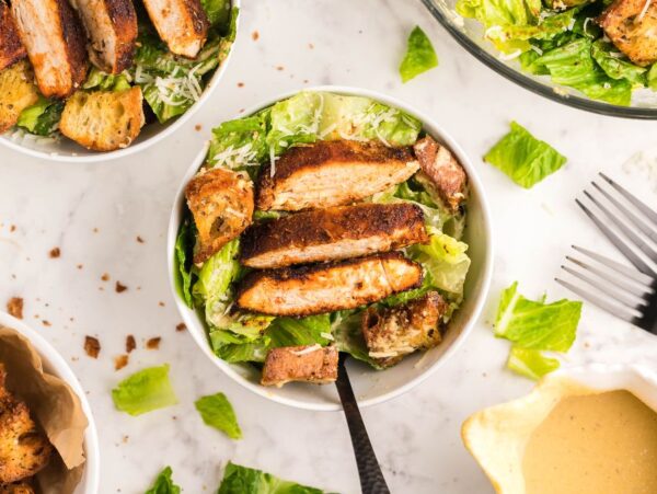 Bowl of Caesar salad topped with air fryer blackened chicken strips and croutons on a white surface, served with the best homemade Caesar dressing.