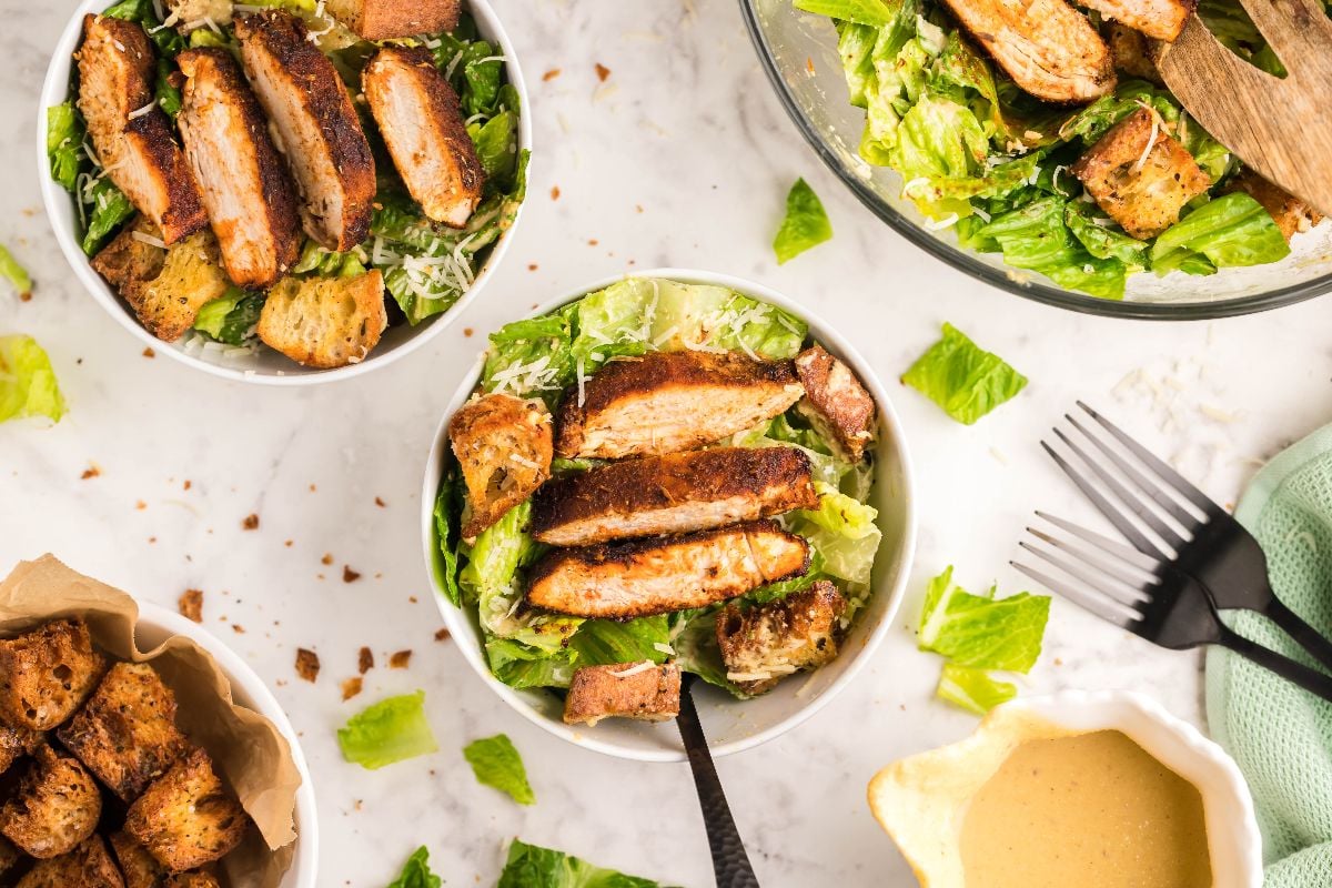 Bowls of Caesar salad topped with grilled chicken and air fryer homemade croutons on a white countertop, with the best homemade Caesar dressing served nearby.