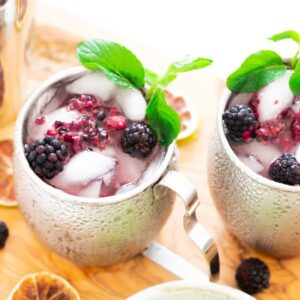 Two silver mugs with ice, blackberries, mint, and a vibrant purple Blackberry Moscow mule sit on a wooden board with dried citrus slices—an inviting cocktail with vodka that's perfect for anyone learning how to make a Moscow mule.